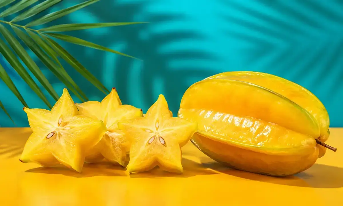 Fruta carambola madura entera y rodajas con forma de estrella sobre fondo amarillo y turquesa — beneficios y peligros de la fruta estrella