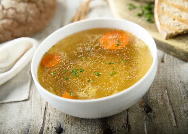 Olla con caldo casero humeante rodeada de verduras frescas como zanahoria y apio
